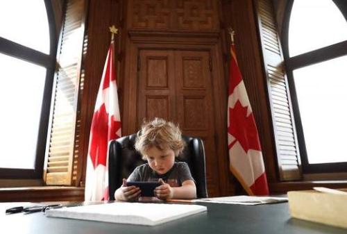 特鲁多儿子坐在爸爸办公椅上看手机。身后是加拿大国旗。